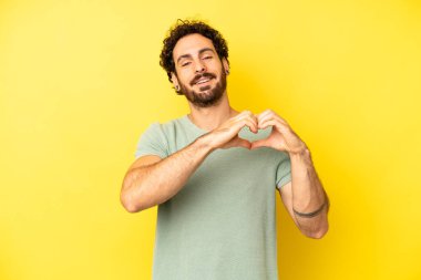 Genç sakallı adam gülümsüyor ve mutlu, sevimli, romantik ve aşık, iki eliyle kalbi şekillendiriyor.