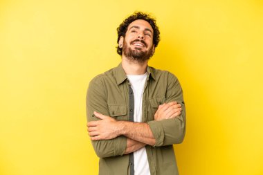 Genç sakallı adam kollarını kavuşturup mutlu bir şekilde gülüyor, rahat, pozitif ve tatmin edici bir pozla.