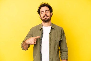 Genç sakallı adam gururlu, kendinden emin ve mutlu görünüyor, gülümsüyor ve kendini işaret ediyor ya da bir numaralı işareti yapıyor.