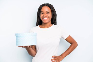 Siyah güzel bir kadın mutlu bir şekilde gülümsüyor, eli kalçasında ve kendinden emin. hediye kutusu konsepti