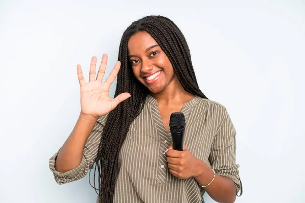 Siyah, güzel bir kadın gülümsüyor ve arkadaş canlısı görünüyor. Beş numarayı gösteriyor. Mikrofonlu şarkıcı