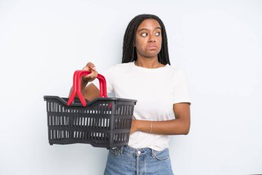 black pretty woman shrugging, feeling confused and uncertain. empty shopping basket