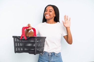 black pretty woman smiling happily, waving hand, welcoming and greeting you. empty shopping basket