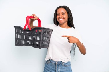 black pretty woman smiling cheerfully, feeling happy and pointing to the side. empty shopping basket