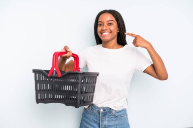 black pretty woman smiling confidently pointing to own broad smile. empty shopping basket