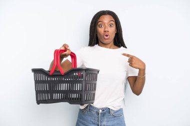 black pretty woman looking shocked and surprised with mouth wide open, pointing to self. empty shopping basket