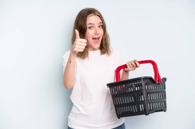 caucasian pretty woman feeling proud,smiling positively with thumbs up. empty shopping basket concept