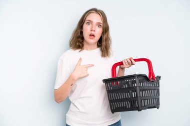 caucasian pretty woman looking shocked and surprised with mouth wide open, pointing to self. empty shopping basket concept