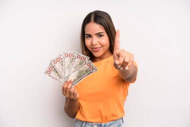 hispanic pretty woman smiling and looking friendly, showing number one. dollar banknotes savings concept