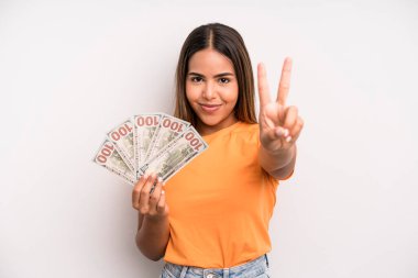 hispanic pretty woman smiling and looking friendly, showing number two. dollar banknotes savings concept