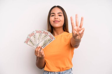hispanic pretty woman smiling and looking friendly, showing number three. dollar banknotes savings concept