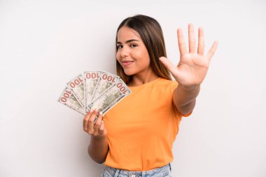 hispanic pretty woman smiling and looking friendly, showing number five. dollar banknotes savings concept
