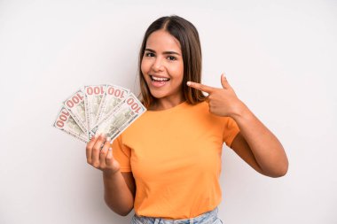 hispanic pretty woman smiling confidently pointing to own broad smile. dollar banknotes savings concept