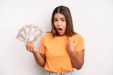 hispanic pretty woman looking shocked and surprised with mouth wide open, pointing to self. dollar banknotes savings concept