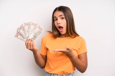 hispanic pretty woman looking surprised and shocked, with jaw dropped holding an object. dollar banknotes savings concept