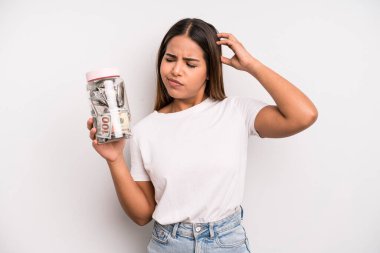 hispanic pretty woman feeling puzzled and confused, scratching head. dollar banknotes savings concept