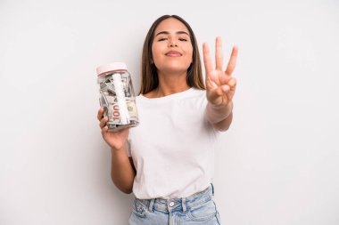 hispanic pretty woman smiling and looking friendly, showing number three. dollar banknotes savings concept