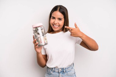 hispanic pretty woman smiling confidently pointing to own broad smile. dollar banknotes savings concept