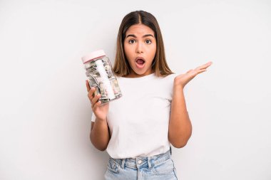 hispanic pretty woman looking surprised and shocked, with jaw dropped holding an object. dollar banknotes savings concept
