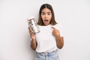 hispanic pretty woman looking shocked and surprised with mouth wide open, pointing to self. dollar banknotes savings concept