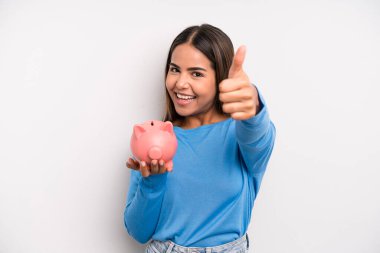İspanyol güzel kadın gururlu, başparmağıyla olumlu gülümsüyor. Domuz kumbarası konsepti
