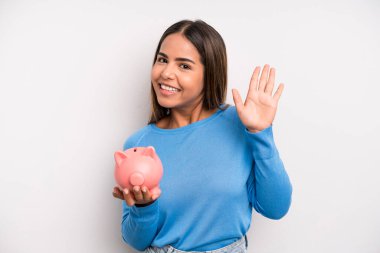 İspanyol güzel kadın mutlu bir şekilde gülümsüyor, el sallıyor, seni selamlıyor ve selamlıyor. Domuz kumbarası konsepti