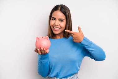Hispanik güzel kadın kendi gülüşünü işaret ederek kendine güvenerek gülümsüyor. Domuz kumbarası konsepti