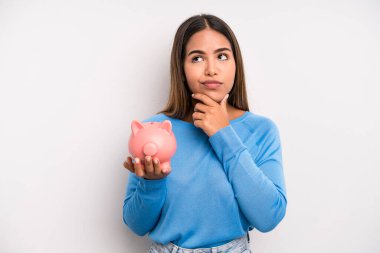 Hispanik güzel kadın düşünüyor, kuşkulu ve şaşkın hissediyor. Domuz kumbarası konsepti