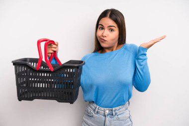 hispanic pretty woman feeling puzzled and confused and doubting. empty supermarket basket concept