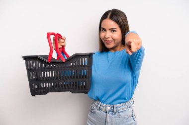 hispanic pretty woman pointing at camera choosing you. empty supermarket basket concept