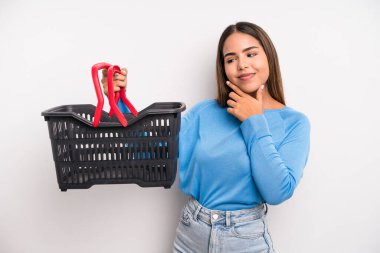 hispanic pretty woman smiling with a happy, confident expression with hand on chin. empty supermarket basket concept