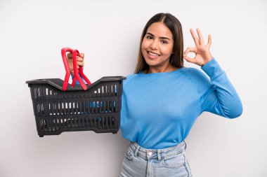 hispanic pretty woman feeling happy, showing approval with okay gesture. empty supermarket basket concept