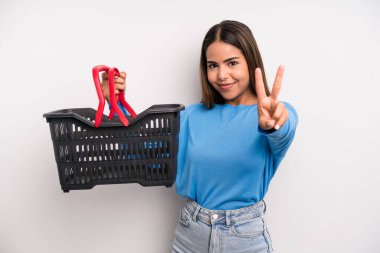 hispanic pretty woman smiling and looking happy, gesturing victory or peace. empty supermarket basket concept