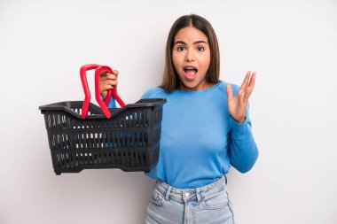 hispanic pretty woman feeling happy and astonished at something unbelievable. empty supermarket basket concept