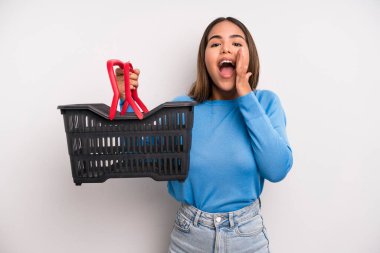 hispanic pretty woman feeling happy,giving a big shout out with hands next to mouth. empty supermarket basket concept