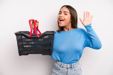 hispanic pretty woman smiling happily, waving hand, welcoming and greeting you. empty supermarket basket concept