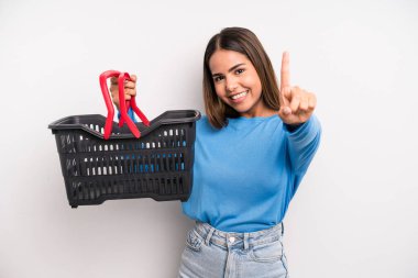 hispanic pretty woman smiling and looking friendly, showing number one. empty supermarket basket concept