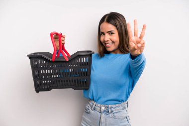 hispanic pretty woman smiling and looking friendly, showing number three. empty supermarket basket concept