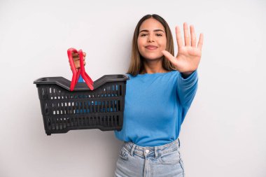 hispanic pretty woman smiling and looking friendly, showing number five. empty supermarket basket concept