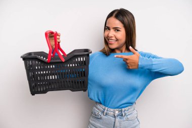 hispanic pretty woman smiling cheerfully, feeling happy and pointing to the side. empty supermarket basket concept
