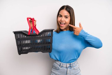 hispanic pretty woman smiling confidently pointing to own broad smile. empty supermarket basket concept