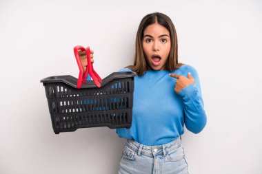 hispanic pretty woman looking shocked and surprised with mouth wide open, pointing to self. empty supermarket basket concept