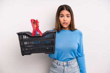hispanic pretty woman looking puzzled and confused. empty supermarket basket concept