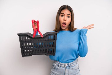 hispanic pretty woman looking surprised and shocked, with jaw dropped holding an object. empty supermarket basket concept
