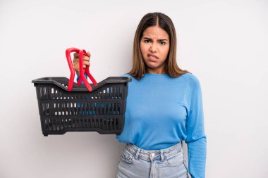 hispanic pretty woman feeling puzzled and confused. empty supermarket basket concept