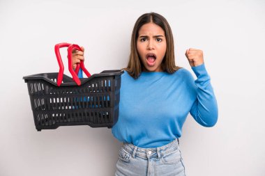 hispanic pretty woman shouting aggressively with an angry expression. empty supermarket basket concept