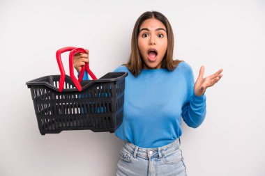 hispanic pretty woman feeling extremely shocked and surprised. empty supermarket basket concept