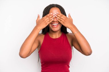 black young adult woman smiling and feeling happy, covering eyes with both hands and waiting for unbelievable surprise