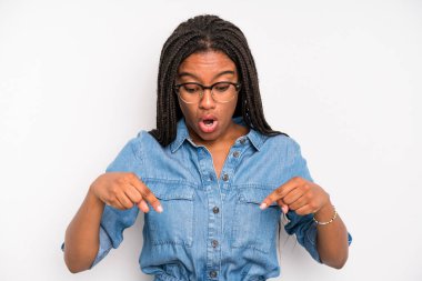 black young adult woman feeling shocked, open-mouthed and amazed, looking and pointing downwards in disbelief and surprise