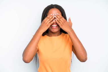 black young adult woman smiling and feeling happy, covering eyes with both hands and waiting for unbelievable surprise